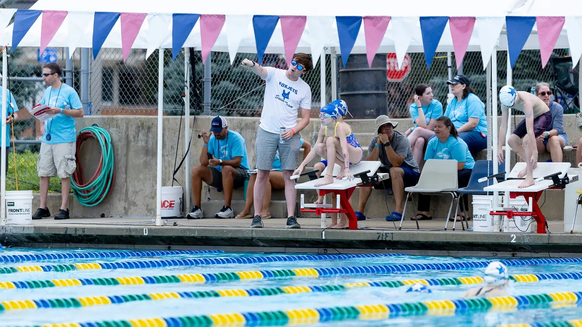 A coach directing swimmers from the poolside at a swim meet, with two swimmers stepping onto the blocks